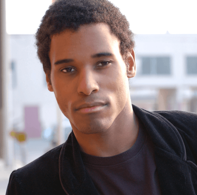 Person wearing a black jacket over a dark shirt, standing outdoors with buildings in the background.